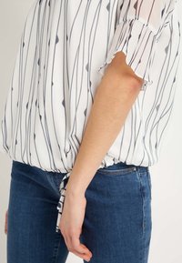 Blouse blanche à rayures verticales bleu marine, avec des manches courtes translucides et un ourlet froncé avec un détail noué, portée avec un jean bleu.