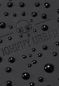 Matowa czarna powierzchnia z wypukłym napisem "JOHNNY URBAN" oraz licznymi błyszczącymi czarnymi kroplami rozsianymi po fakturze.
