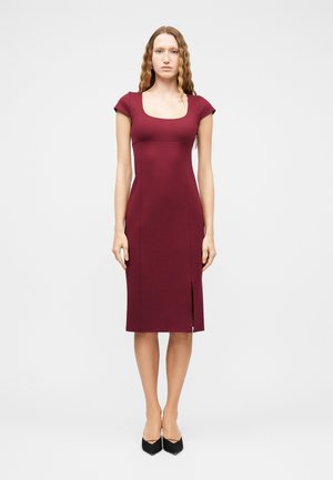 Donna con capelli mossi che indossa un abito aderente bordeaux al ginocchio con maniche a copertura delle spalle e una spaccatura frontale, abbinato a tacchi neri con punta appuntita.