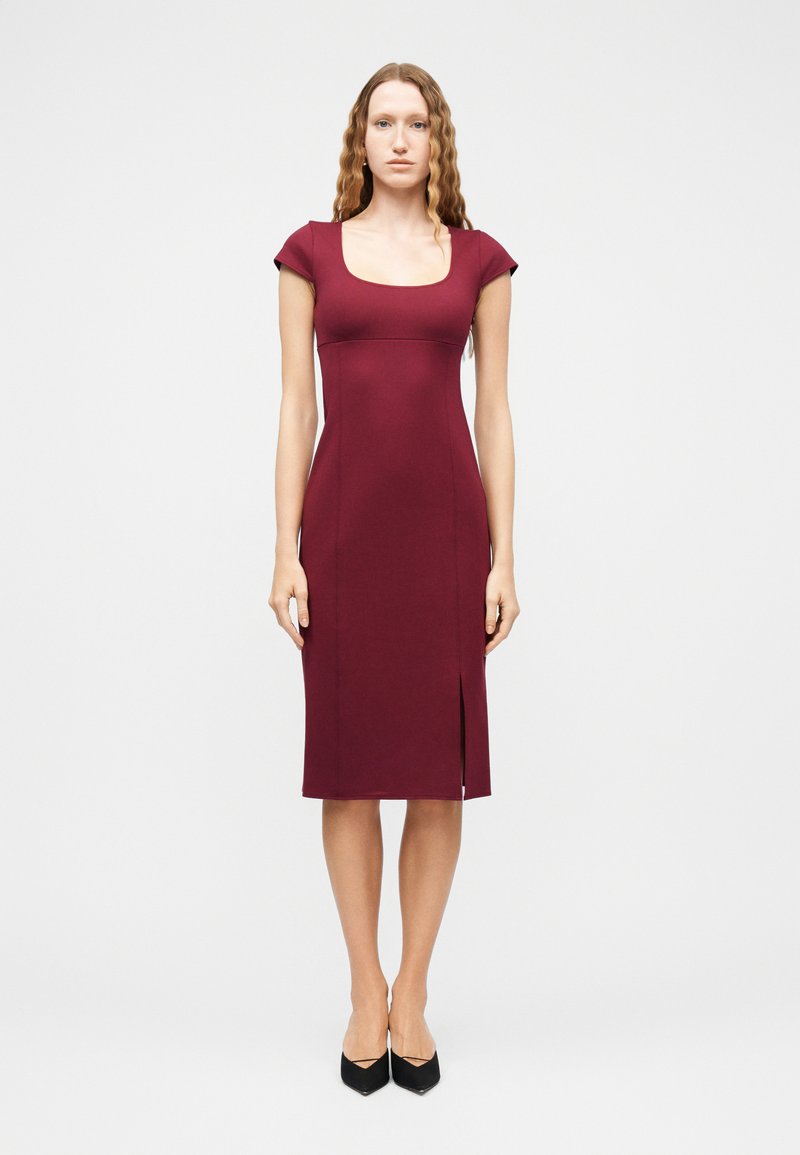 Γυναίκα με κυματιστά μαλλιά που φοράει εφαρμοστό μπορντό φόρεμα μέχρι το γόνατο με κοντά μανίκια και μπροστινή σχισμή, σετ με μαύρες μυτερές γόβες.