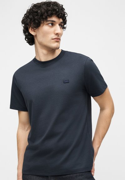 Νέος με σγουρά μαλλιά που φοράει ένα απλό σκούρο μπλε μπλουζάκι με ένα μικρό κεντημένο λογότυπο στο στήθος, κοιτάζοντας προς τα πλάγια.