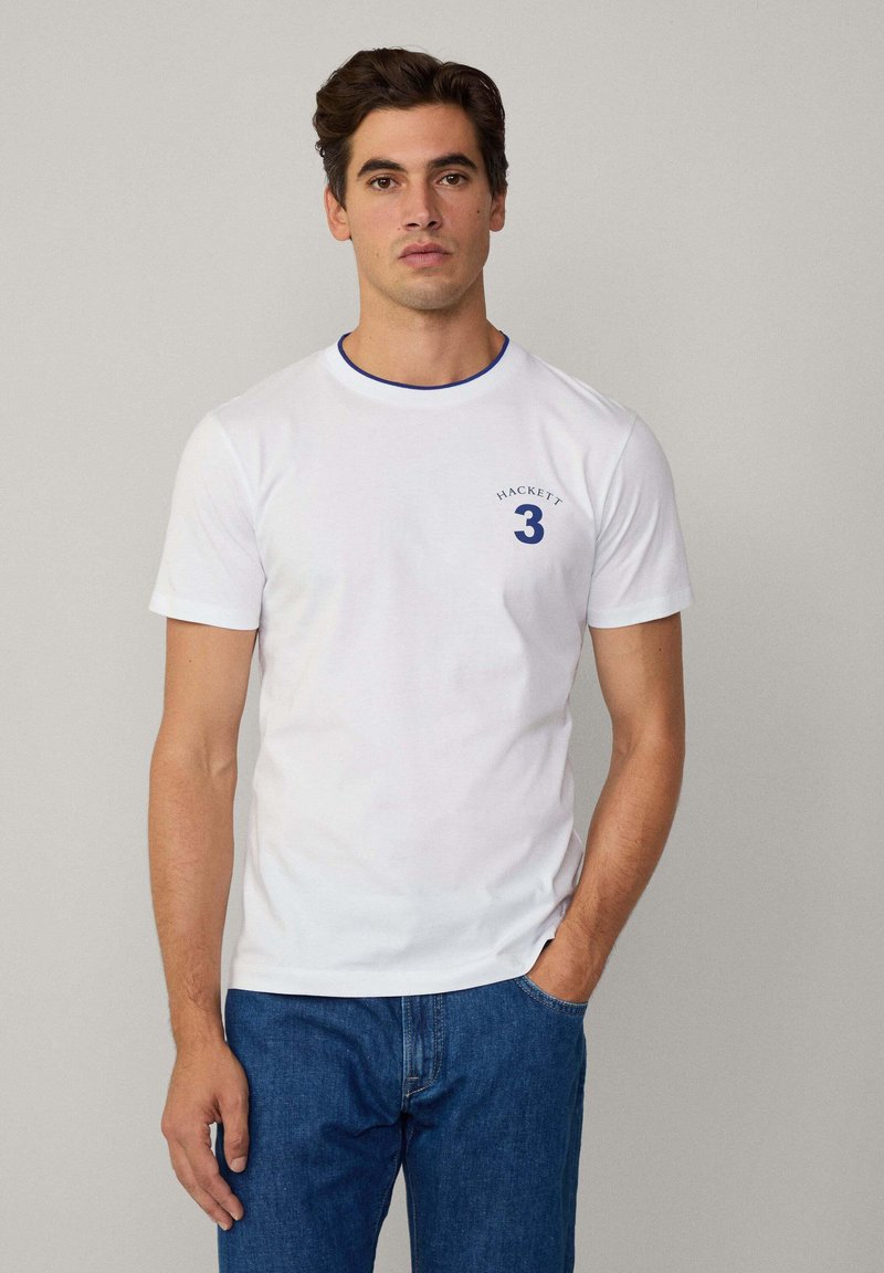 Hvit t-skjorte med korte ermer og blå rund hals, med en sirkulær marineblå print som har "HACKETT 3" på venstre bryst. Kombinert med blå jeans.