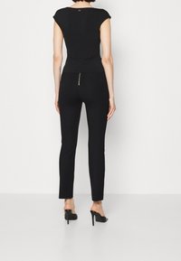 Top negro ajustado con mangas cortas y detalle de torsión en la parte trasera, combinado con pantalones negros ajustados con cremallera en la parte trasera. Llevado con tacones altos negros.
