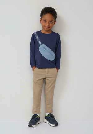 Marineblauwe long-sleeve shirt met een blauwe heuptas, beige broek en donkere sneakers met neonaccenten, staande tegen een neutrale achtergrond.
