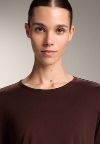 Nuori henkilö, jolla on ruskeat silmät ja taakse kammatut hiukset, pukeutuneena tummanruskeaan pyöreäkauluspaitaan neutraalia taustaa vasten.