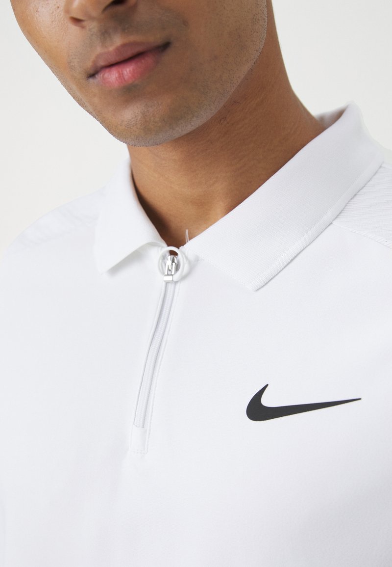 Biała koszulka polo Nike z zamkiem błyskawicznym, eleganckim kołnierzykiem, teksturowanym akcentem na ramieniu oraz wyraźnym czarnym logo po lewej stronie klatki piersiowej.