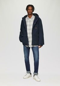 Marine parka jas met een capuchon, knoopflapzakken en een grijs geruite overhemd eronder, gedragen met blauwe spijkerbroek en witte sneakers.