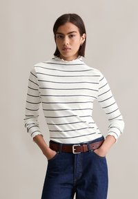 Haut à manches longues à rayures blanches avec un col haut et une bordure volante, associé à un pantalon en denim bleu foncé et une ceinture en cuir marron.