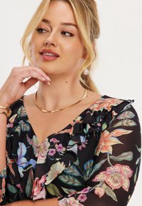 Bloes met bloemenprint van zwarte stof met ruches, met levendige bloemen in verschillende kleuren. Gouden ketting en pareloorbellen maken de look compleet.