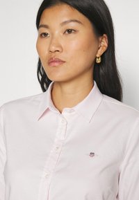 Woman with long dark hair wearing a light pink button-up shirt with a small embroidered logo and gold hoop earrings.