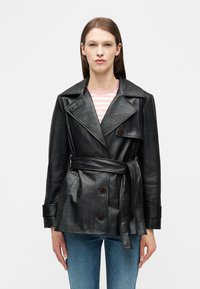 Γυναίκα που φοράει μαύρο δερμάτινο μπουφάν με ζώνη πάνω από ένα ροζ και λευκό ριγέ πουκάμισο και μπλε τζιν, στέκεται μπροστά σε ένα απλό ανοιχτό φόντο.