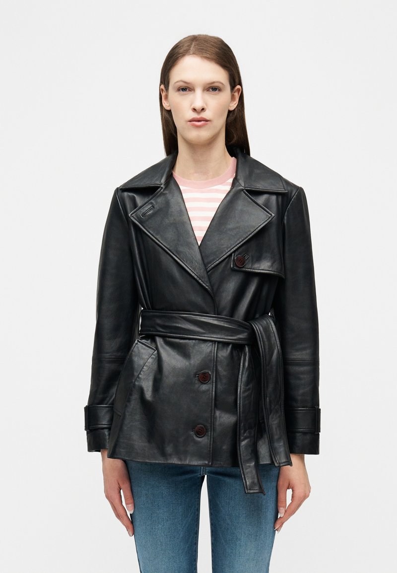 Γυναίκα που φοράει μαύρο δερμάτινο μπουφάν με ζώνη πάνω από ένα ροζ και λευκό ριγέ πουκά μισο και μπλε τζιν, στέκεται μπροστά σε ένα απλό ανοιχτό φόντο.