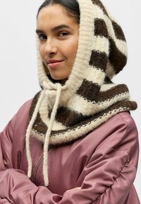 Écharpe à capuche en maille à motifs crème et marron avec une finition douce et texturée ; portée par-dessus une veste bomber rose pâle.