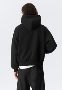 Svart hettegenser med cropped design, ribbet kant og senkede skuldre. Myk tekstur og minimalistisk stil. Brukt med svarte jeans.