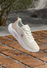 Pantof de sport bej deschis, cu un material din plasă, o talpă albă și un logo roșu. Are un design curbat și accente texturate pe toc.