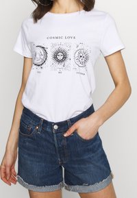 Vit bomullströja med svart grafiskt tryck som har himmelska motiv och texten "COSMIC LOVE", tillsammans med blå denimshorts med fransad nederkant.