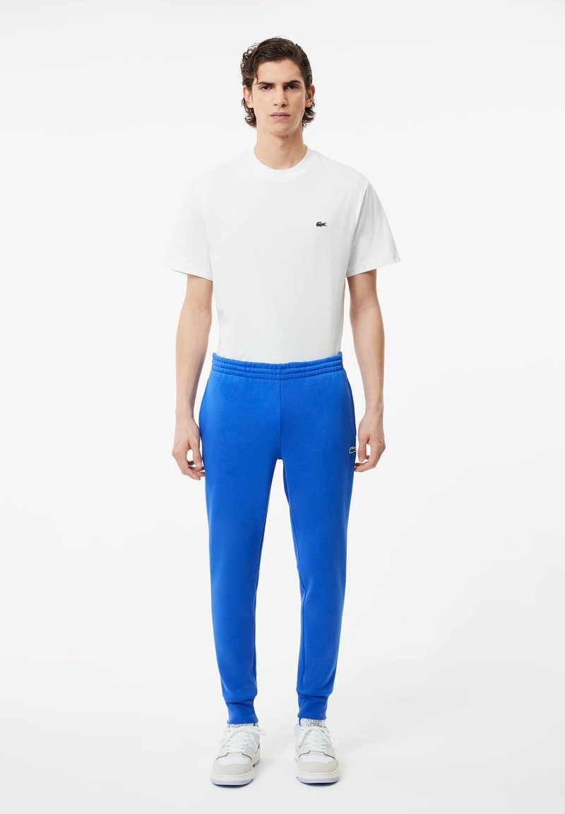 Biała bawełniana koszulka z małym zielonym logo, w połączeniu z niebieskimi spodniami dresowymi z elastycznym pasem i mankietami na kostkach. Czarne sneakersy dopełniają wygląd.