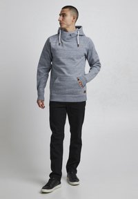 Sudadera de textura gris con un gran cuello, cordones blancos, bolsillo frontal y parche de piel marrón. Combinada con pantalones y calzado negros.