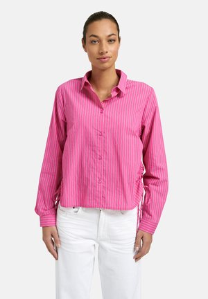 Frau trägt ein langärmliges rosa Hemd mit weißen Streifen, vorne geknöpft, und weiße Hosen, steht vor einem schlichten Hintergrund.