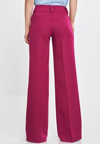 Osoba w wysokich szerokich nogawkach magentowych spodni z tylnymi kieszeniami, stojąca na tle jednolitego białego tła.