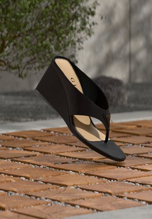 Black wedge sandal with thong strap and gold triangular logo emblem, displayed above textured brick ground with greenery and wall in background.
