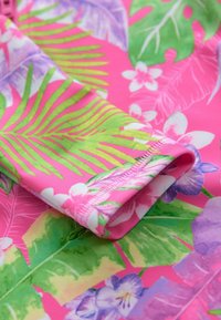 Rosa tropiskt mönstrad tyg med stora gröna löv och lila blommor. Designen inkluderar en rullad manschett med sydda kanter.