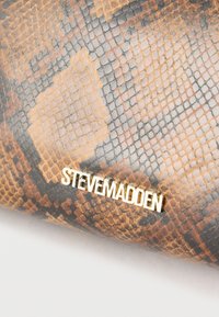 Καφέ και μαύρη τσάντα με μοτίβο φίδι και χρυσό λογότυπο "Steven Madden" σε λευκό φόντο.