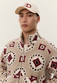 Mlad moški, oblečen v bež flis jakno z rdečimi in rjavimi geometrijskimi vzorci ter krem baseball kapo z rdečimi vezenimi črkami.