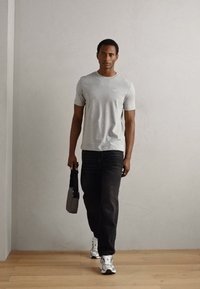 Tricou gri cu mânecă scurtă, blugi negri, adidași gri. Ținând o geantă gri de umăr. Stând împotriva unei pereți deschise la culoare, pe un podea din lemn.