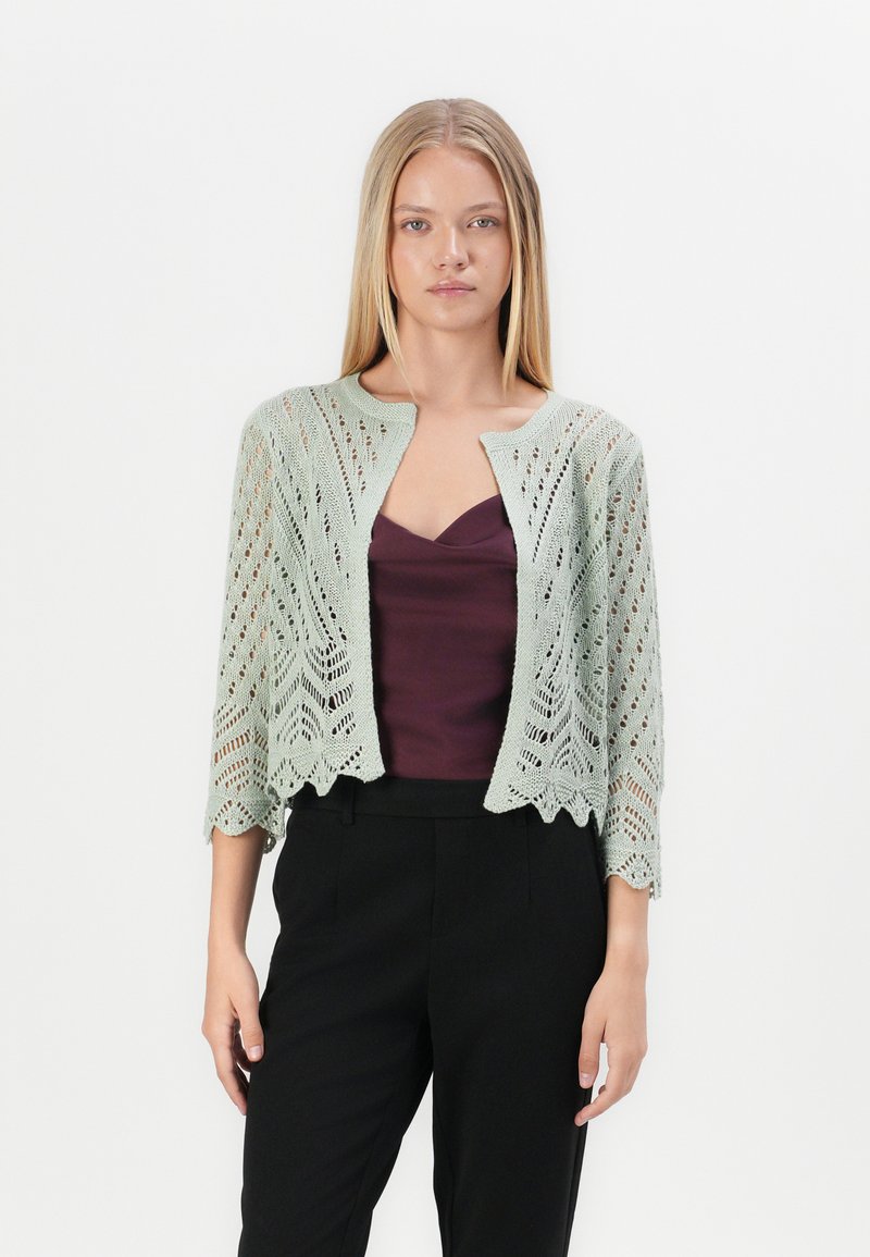 Gebreide cardigan in lichtgroen, met een gegolfde zoom, open voorkant en een ingewikkeld kanten patroon. Gedragen over een diep paarse top en zwarte broek.