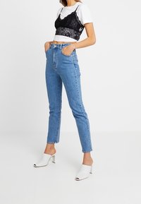 Top court en dentelle noire superposé sur un T-shirt blanc, associé à un jean en denim bleu clair taille haute et des mules blanches à talons ouvertes à l'avant.