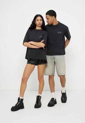 Ung kvinde iført sort oversized t-shirt, sorte shorts og støvler står med armene over kors ved siden af en mand i sort t-shirt, denimshorts og loafers.