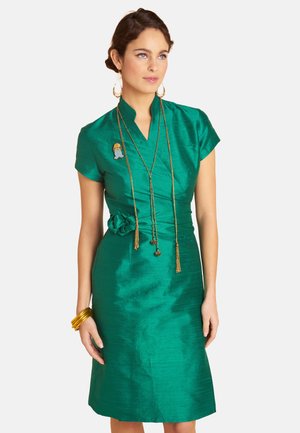 Woman wearing a shiny green knee-length dress with short sleeves, gold jewelry, and a flower detail on the waist, standing against a plain background.