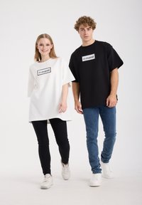 Oversized T-shirts in wit en zwart katoen. Beide hebben een rechthoekig logo met tekst. Model draagt zwarte spijkerbroek en witte sneakers.