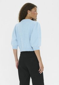 Vrouw met krullend haar, die een lichtblauwe, fuzzy, cropped trui en hoog opgesneden zwarte broek draagt, zijwaarts gekeerd tegen een witte achtergrond.