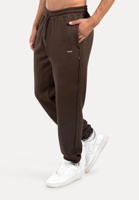 Brązowe spodnie dresowe z elastycznym pasem, bocznymi kieszeniami i zwężanymi mankietami, w połączeniu z białymi butami sportowymi. Materiał wydaje się miękki i o fakturze.