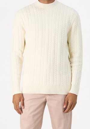 Homme portant un pull en maille torsadée crème et un pantalon beige clair, debout les bras détendus le long du corps.