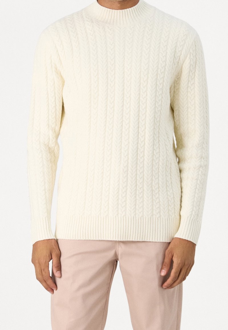 Homme portant un pull en maille torsadée crème et un pantalon beige clair, debout les bras détendus le long du corps.