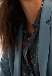 Lichtblauwe blazer van lichtgewicht stof, gecombineerd met een donker overhemd met een bloemenpatroon in groene en roze tinten, met knopen op de kraag.