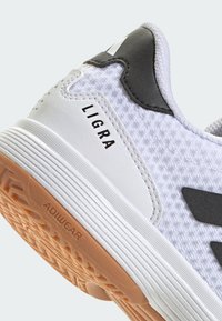 Baskets blanches avec tige texturée, accents noirs et semelle en caoutchouc. Présente le logo "LIGRA" et "ADIWEAR" sur le côté et la semelle.