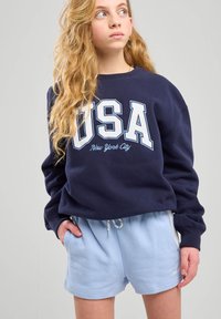 Felpa blu navy con testo "USA" e "New York City" in bianco, abbinata a pantaloni corti azzurri chiari con vita elasticizzata e tasche.