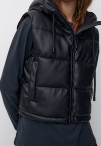 Veste sans manches matelassée noire avec capuche, présentant une texture élégante en cuir synthétique, fermeture éclair et cordons ajustables.