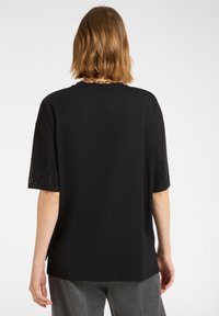 Czarna koszulka wykonana z miękkiego materiału, z krótkimi rękawami ozdobionymi cyrkoniami. Okrągły dekolt i luźny krój.