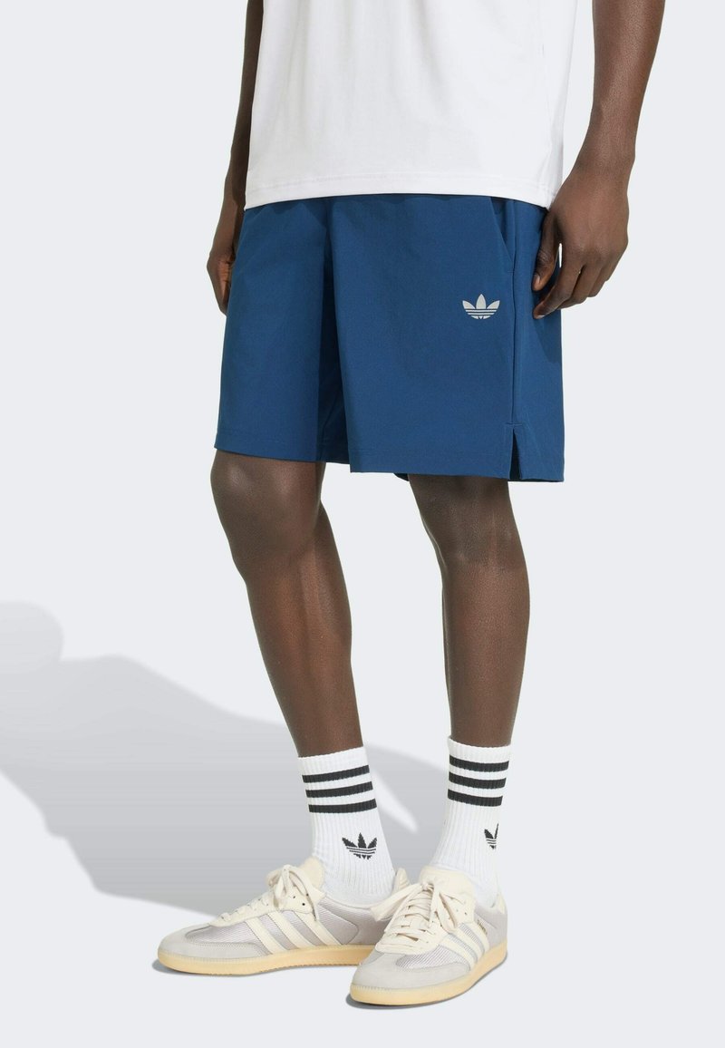 Mann trägt weiße Adidas-Sneaker und Socken mit schwarzen Streifen, blaue Adidas-Shorts und weißes Shirt, steht vor hellem Hintergrund.
