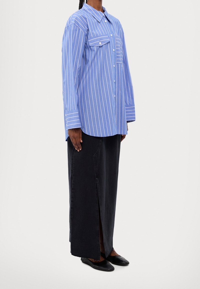 Personne portant une chemise à manches longues rayée verticalement bleu et blanc, une jupe longue noire avec une fente à l'avant et des chaussures plates noires, debout de côté.