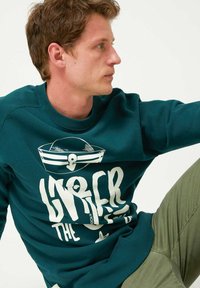 Joven con sudadera color teal y un sombrero de marinero blanco con un gráfico de ancla, vestido con pantalones verde oliva, mirando de lado ante un fondo simple.