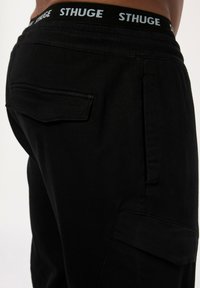 Schwarze Cargo-Hosen mit schlankem Design, elastischem Bund mit "STHUGE"-Logo und einer aufgesetzten Tasche auf der Rückseite. Glatte Textur.