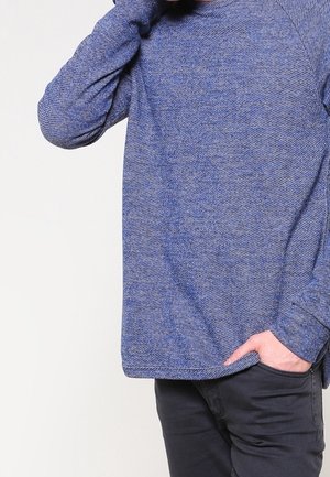 Mand iført en tekstureret blå langærmet sweater med den ene hånd, der rører ved nakken, og den anden i lommen på sorte bukser.