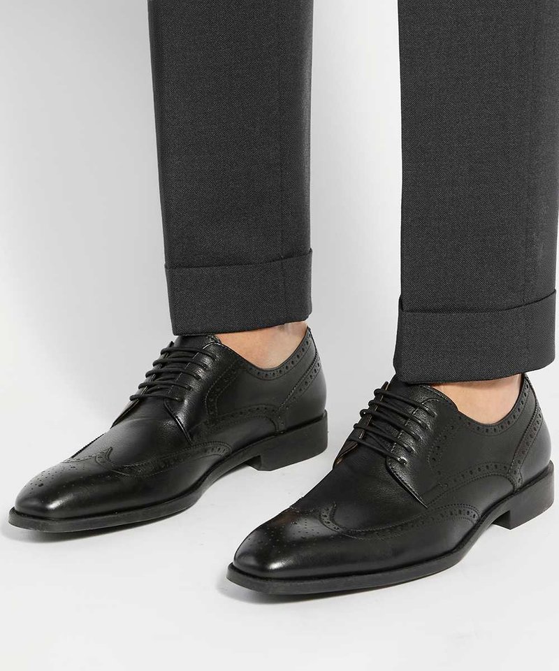 Chaussures de ville brogue en cuir noir portées avec un pantalon gris foncé à revers sur fond blanc.