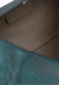 Interno di una borsa in pelle teal con una fodera morbida e di un marrone chiaro. Presenta due scomparti e una tasca rettangolare a soffietto. Dettagli di cucitura visibili.
